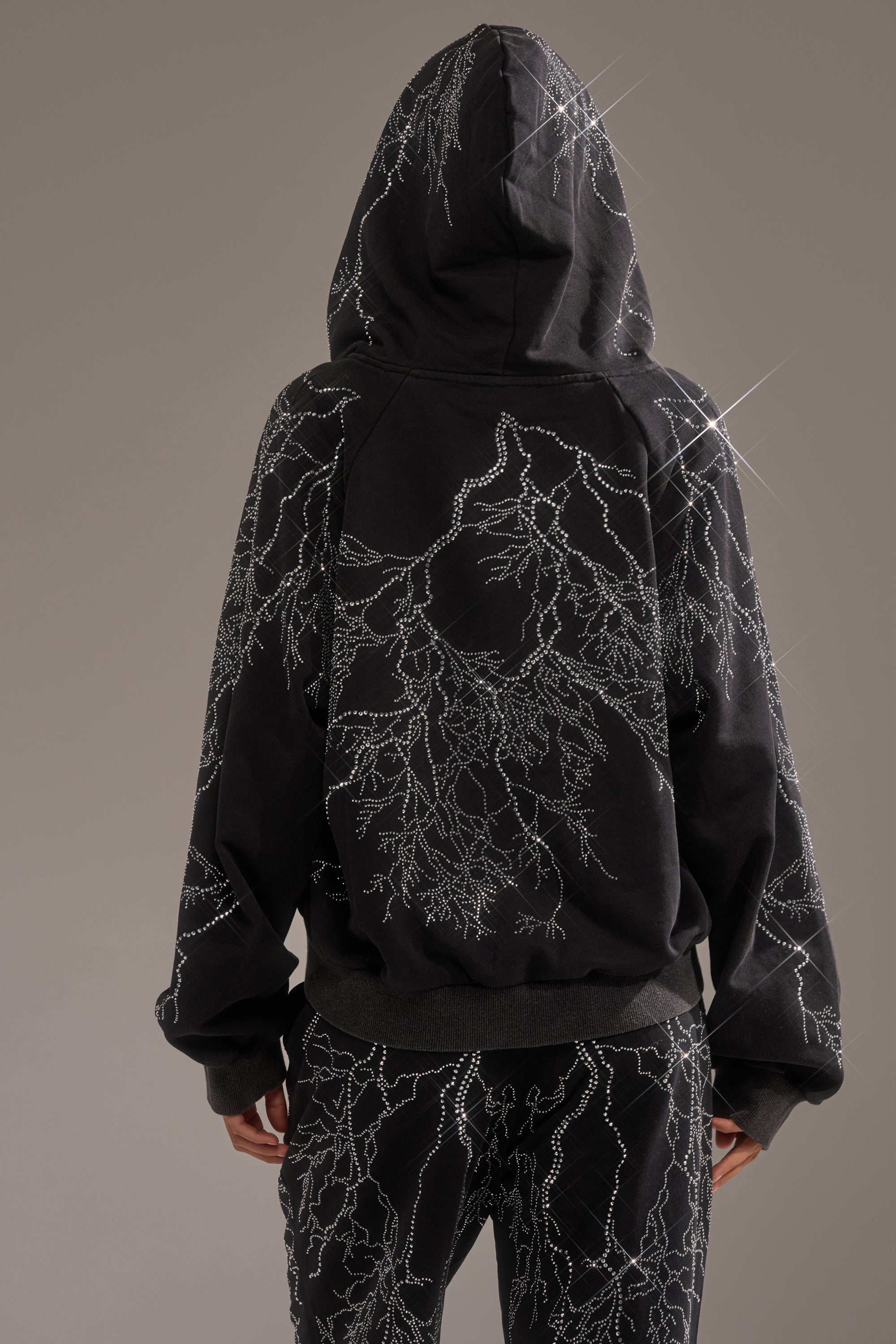 Someone wearing the ELECTRIC LOVE RHINESTONE ZIP UP SWEATSHIRT, detailed with sparkling white lightning-like rhinestone patterns, stands with their back to the camera against a neutral background. 3