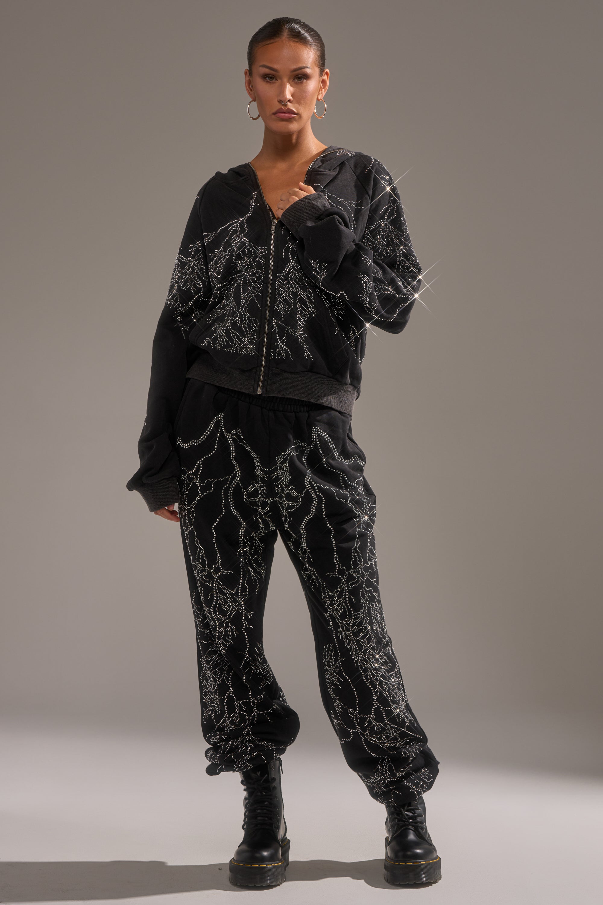 A woman stands confidently in the ELECTRIC LOVE RHINESTONE ZIP UP SWEATSHIRT and matching joggers with bold white lightning patterns, chunky black boots, hoop earrings, and slicked-back hair, posing against a plain gray background. 5