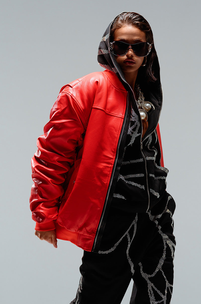 Person posing confidently in a red leather jacket over an AKIRA Label Spider Sequin Detail Zip Up Hoodie with silver patterns, wearing black pants with matching rhinestone detailing. They also have oversized black sunglasses and an elaborate silver necklace, set against a plain background, showcasing a cozy glam ensemble.