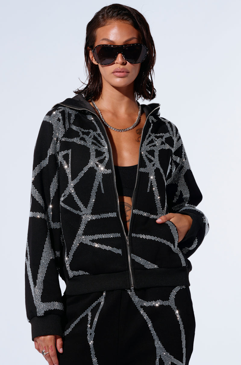 Person wearing large black sunglasses, a cozy glam ensemble featuring the AKIRA Label Spider Sequin Detail Zip Up Hoodie with rhinestone detailing and matching black pants. The individual has their hands in the jacket pockets and is standing against a light-colored background.
