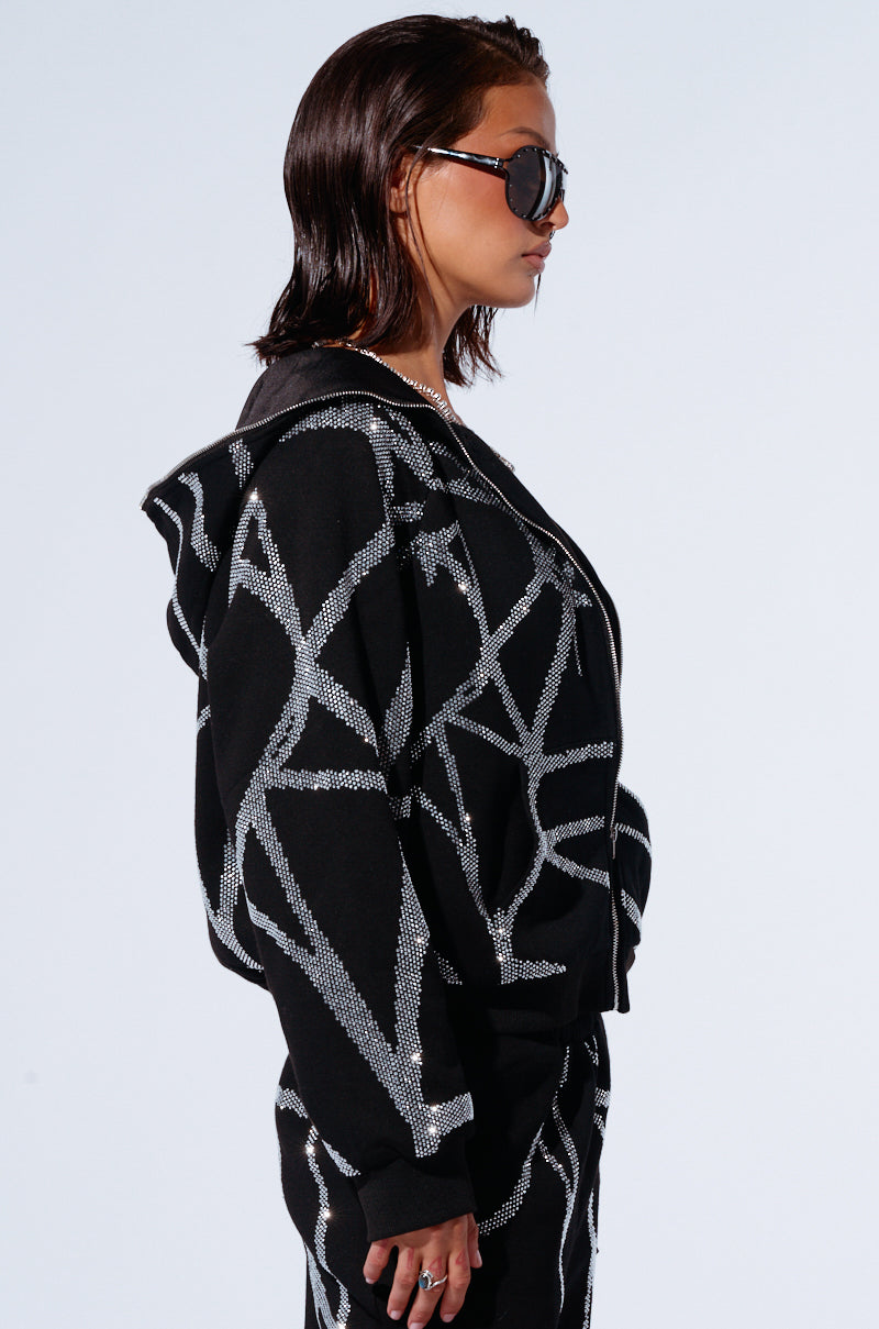 A person with short, slicked-back hair is shown in a side profile wearing dark sunglasses and an AKIRA SPIDER SEQUIN DETAIL ZIP UP HOODIE. The black outfit features geometric silver patterns and rhinestone detailing, creating a cozy yet glamorous ensemble. The background is plain and light-colored.