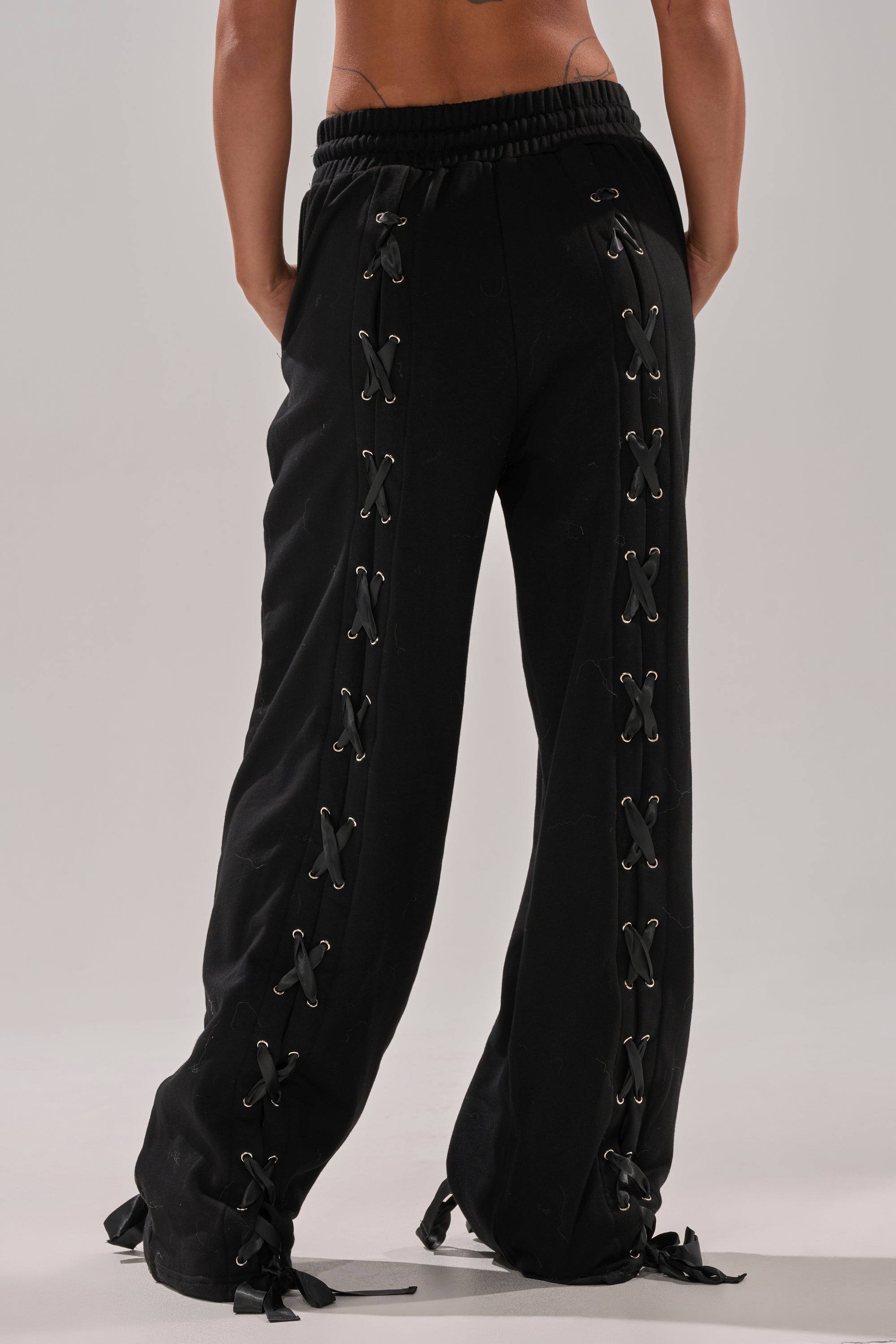 A person stands facing away in ALL LACED UP STRAIGHT-LEG SWEATPANTS, featuring black crisscross lace-up details along the sides and hems. These relaxed fit sweatpants have an elastic waistband. The background is plain and neutral. 4
