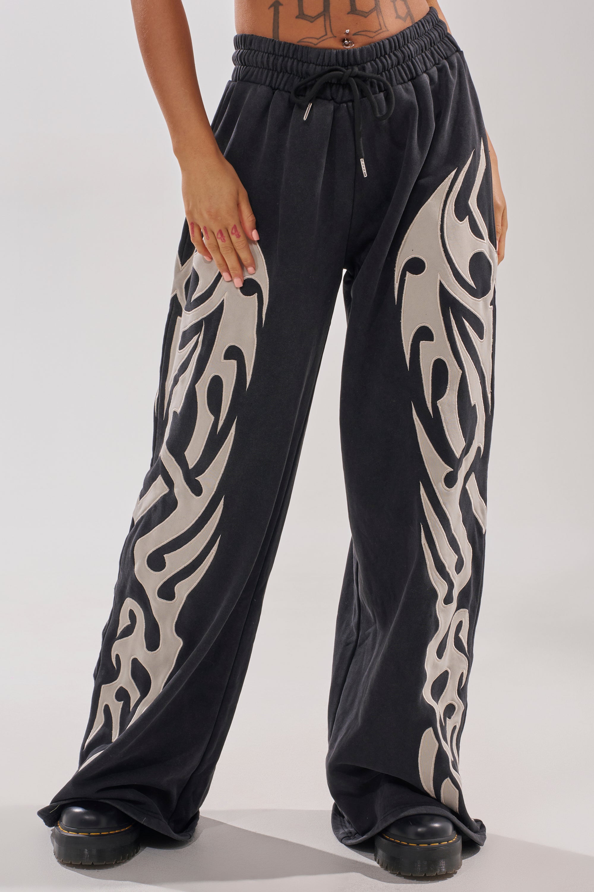 A person wears the ON FIRE WIDE LEG JOGGER, featuring bold, light tribal patterns on the legs. Their midriff and tattooed lower abdomen are visible, paired with black shoes.
