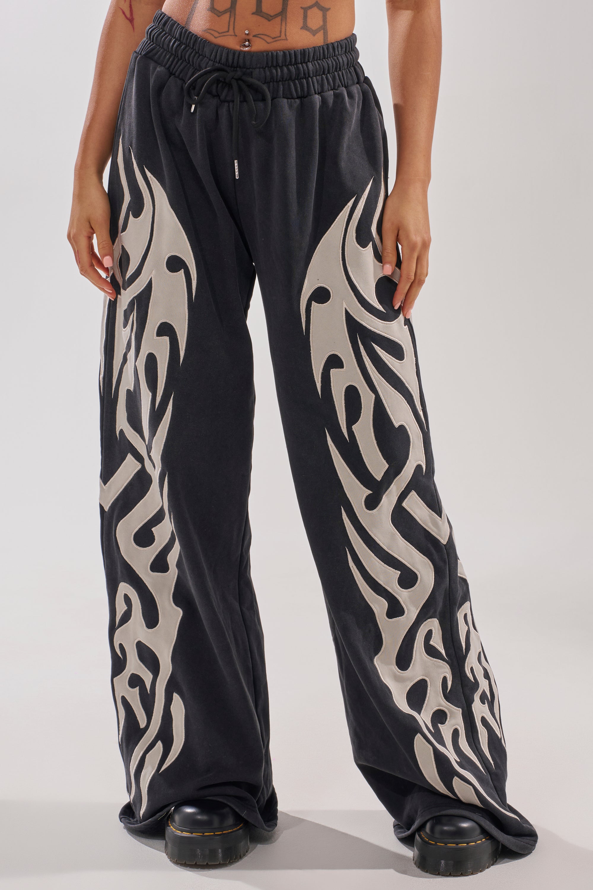 Someone is shown from the waist down wearing ON FIRE WIDE LEG JOGGER pants featuring bold beige flame graphics on both legs, paired with black chunky shoes.