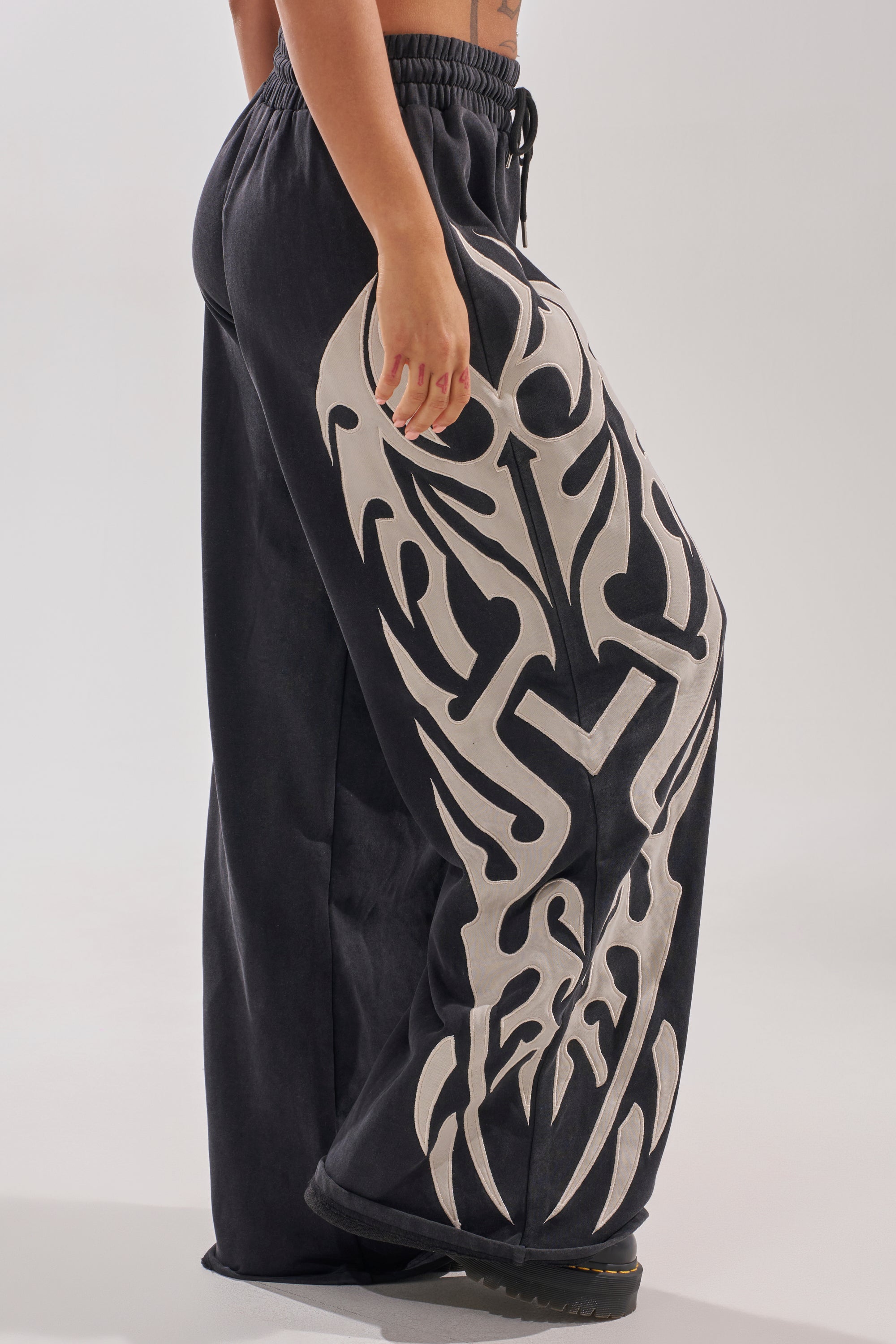 A person wearing ON FIRE WIDE LEG JOGGER pants with a bold white tribal design on the side, standing sideways with one arm relaxed by their side.