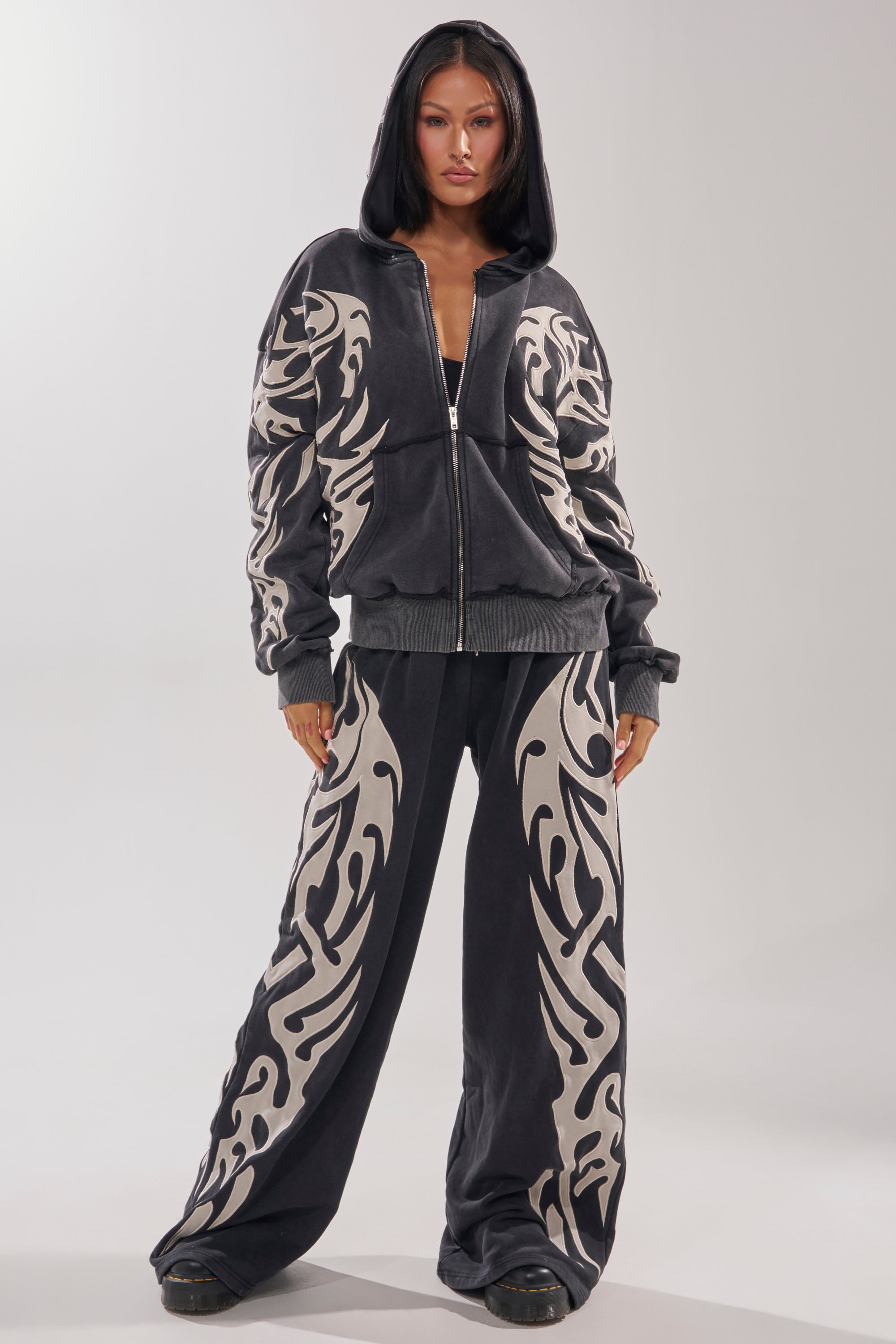 A woman stands forward-facing in the ON FIRE WIDE LEG JOGGER—a black zip-up hoodie and wide leg jogger pants featuring bold cream tribal patterns—her cotton-blend loungewear contrasting her dark hair against a light background.