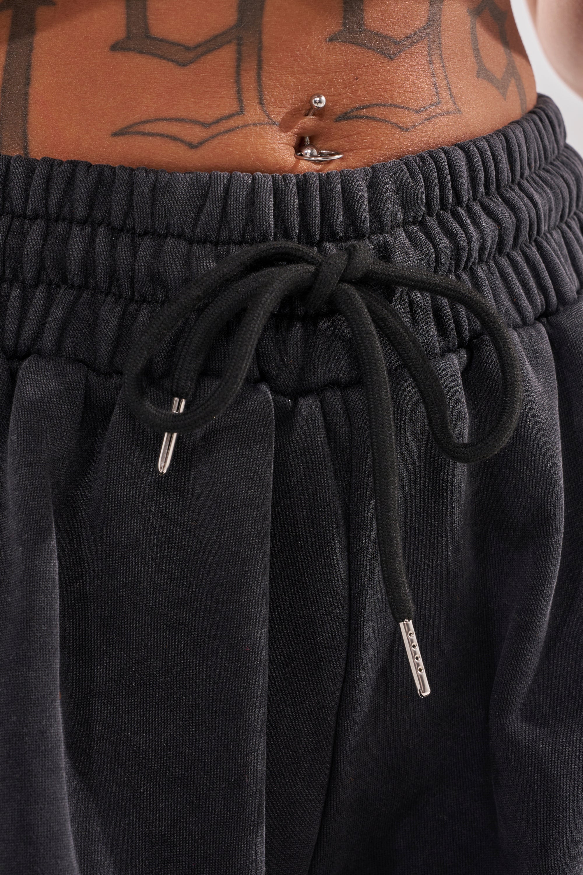 Close-up of a person’s midsection wearing the ON FIRE WIDE LEG JOGGER, black cotton-blend knit sweatpants, with a belly button piercing and part of a geometric tattoo visible above the waistband.