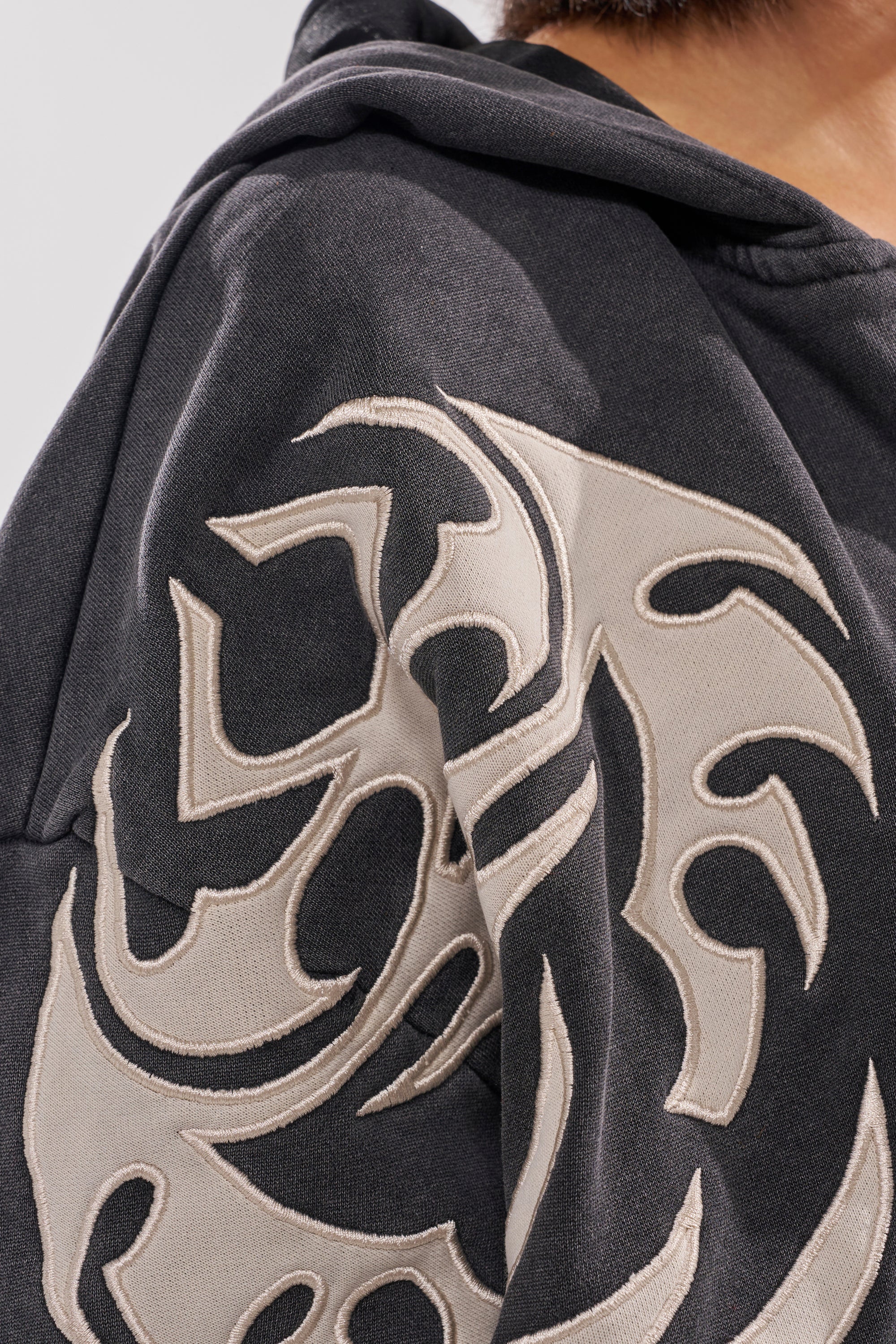 Close-up of a person wearing the ON FIRE ZIP UP SWEATSHIRT, an oversized dark hoodie with a large light flame embroidery on the sleeve and shoulder. The person's face is not shown. 9