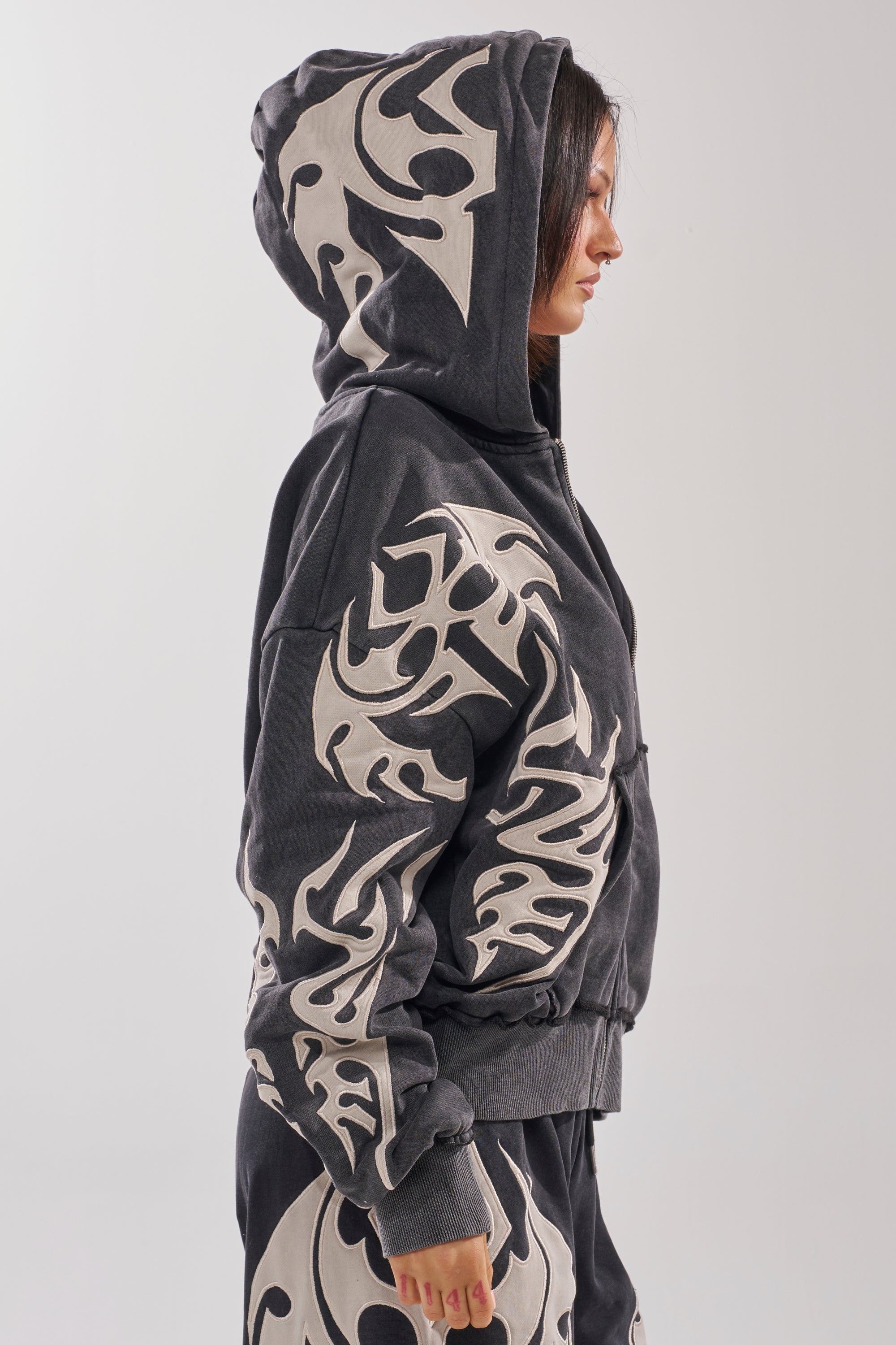 A woman in profile against a plain light background wears the ON FIRE ZIP UP SWEATSHIRT, a black oversized piece featuring bold beige tribal patterns and an embroidered flame design. 4