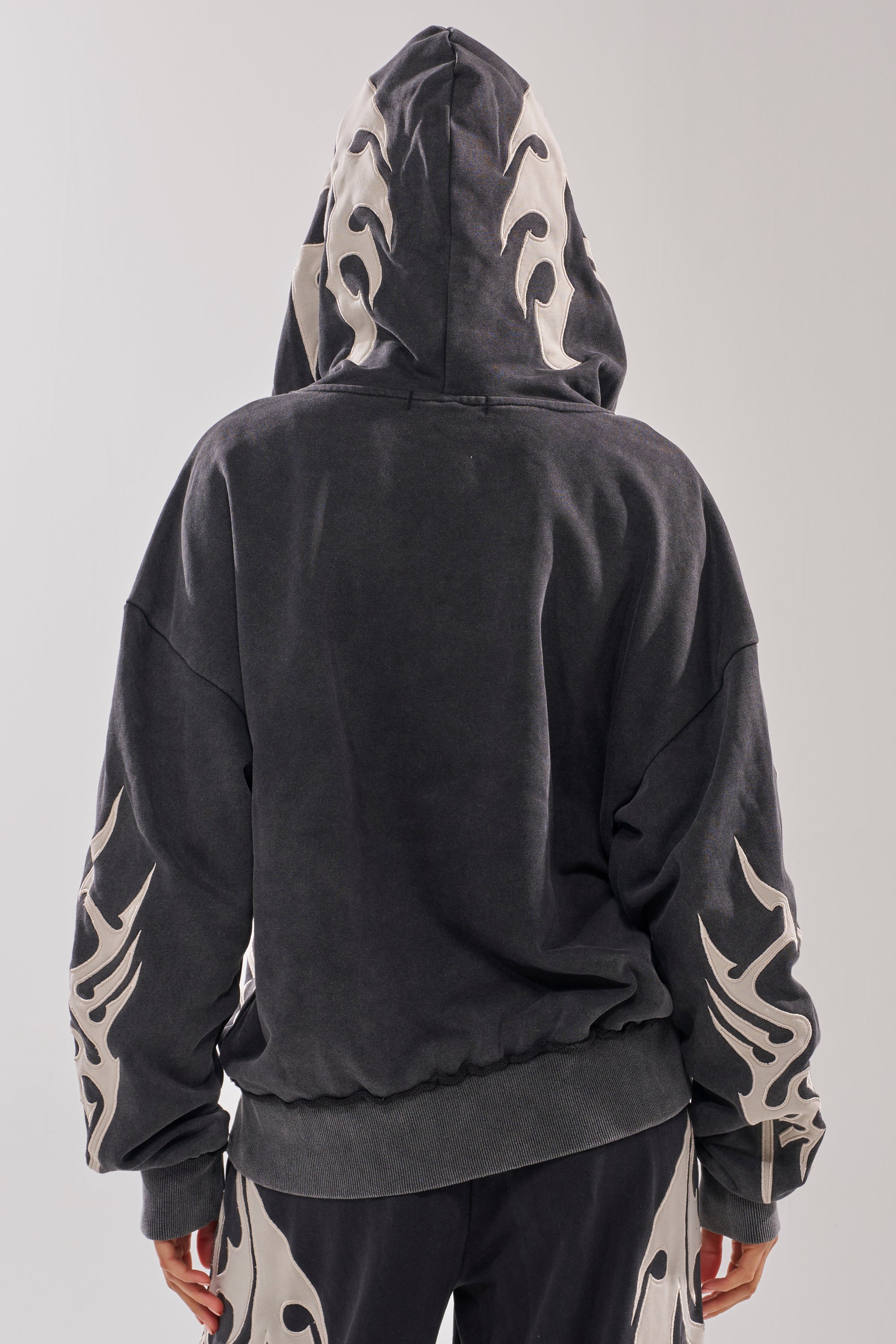 Someone wears the ON FIRE ZIP UP SWEATSHIRT, featuring white flame designs on the hood and sleeves, seen from behind. The outfit is paired with matching pants with similar flame patterns, hands resting at their sides. 5