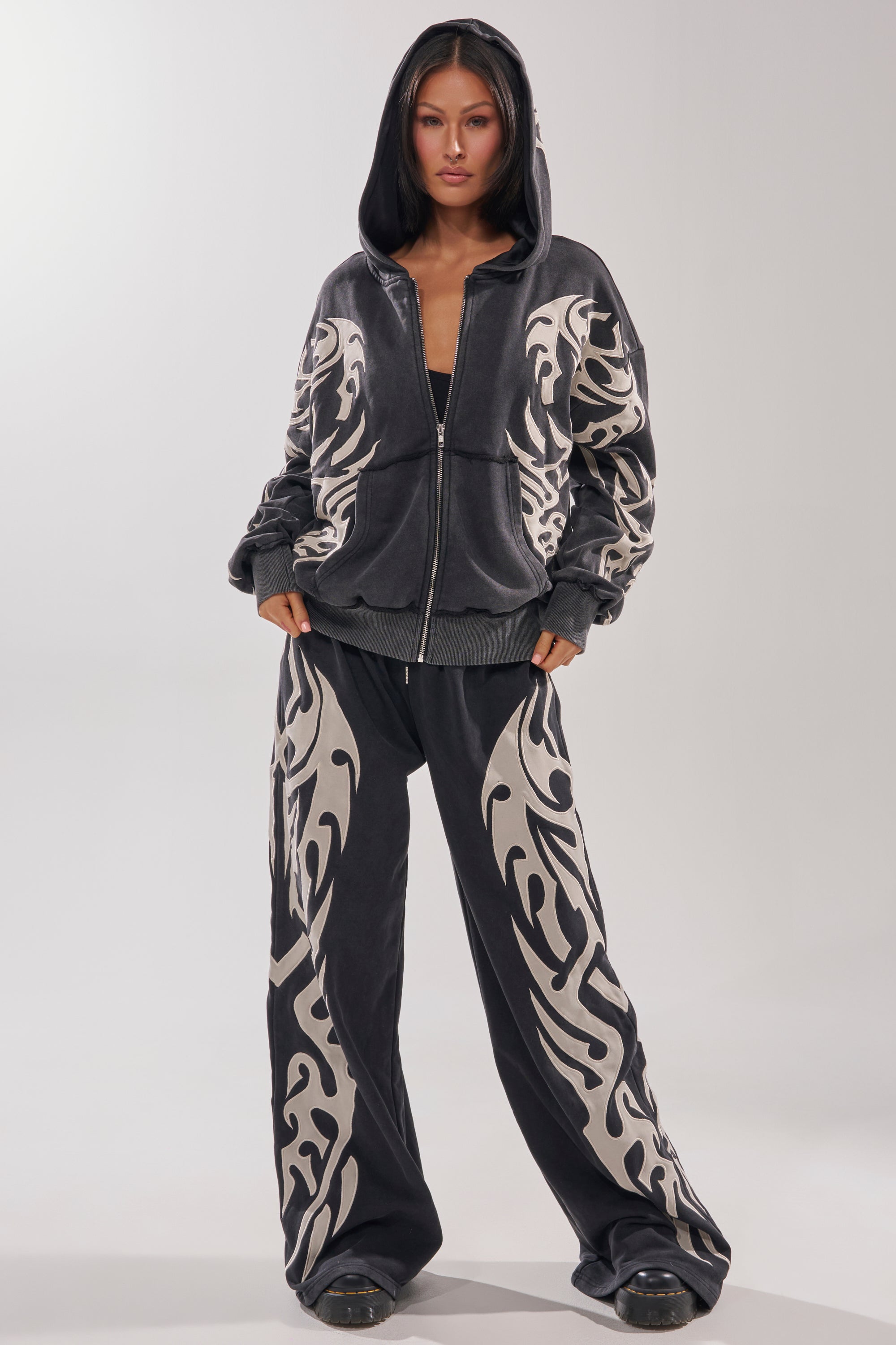 A woman faces forward in the ON FIRE ZIP UP SWEATSHIRT—an oversized black velour tracksuit with bold white tribal patterns and an embroidered flame motif. She wears the hood up, hands in her pockets, against a plain light backdrop. 6
