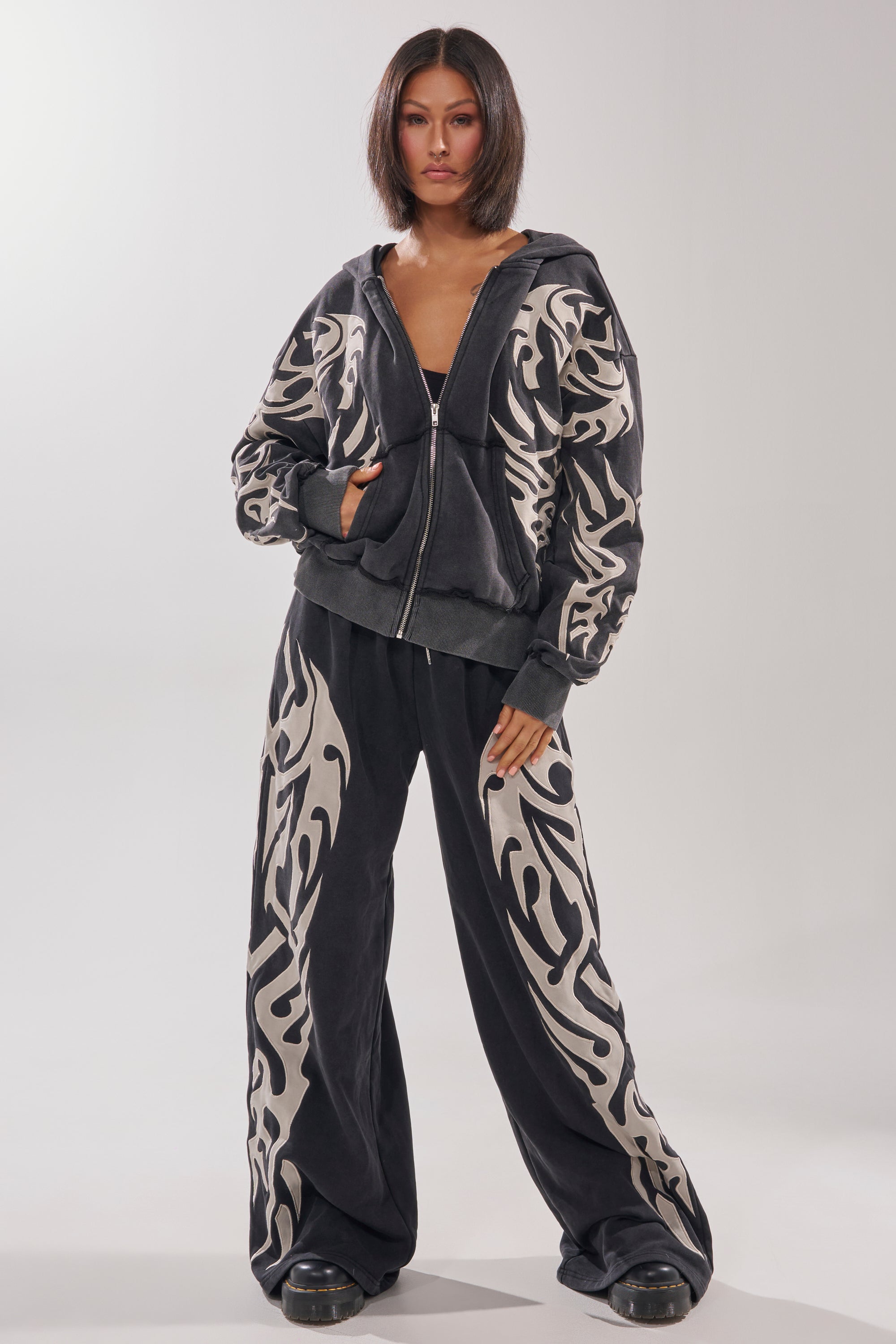A person stands facing forward in an oversized ON FIRE ZIP UP SWEATSHIRT and matching wide-leg pants with bold white tribal patterns. The hoodie has an embroidered flame design. They have straight shoulder-length dark hair and wear black shoes. 7