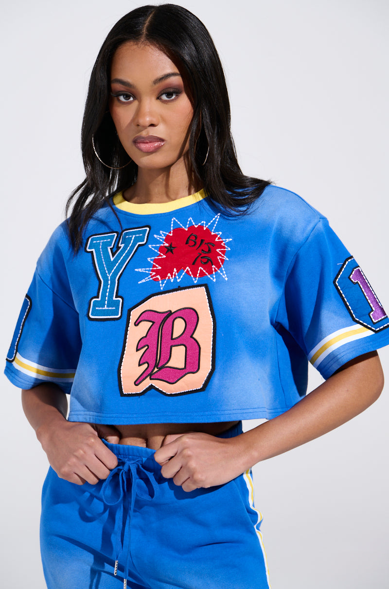 A woman with long black hair wears the THREE ONE TWO CROP TEE by AKIRA Label, a vibrant blue sports jersey embellished with colorful patches of letters and symbols. She accessorizes with hoop earrings and displays a confident look against a plain backdrop.