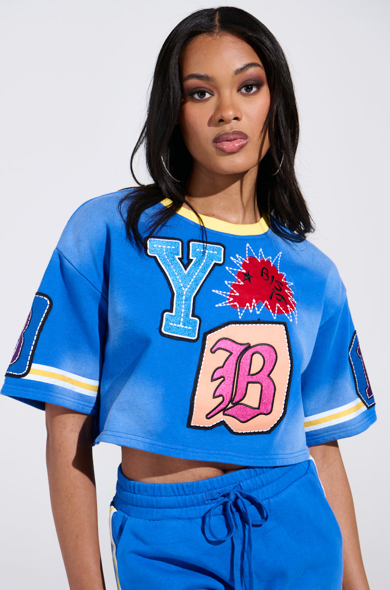A Chicagoan with long hair wears the AKIRA Label's THREE ONE TWO CROP TEE, a blue cropped sweatshirt adorned with colorful patches such as "Y," "B," and a bold red burst design. They finish their look with matching blue pants.