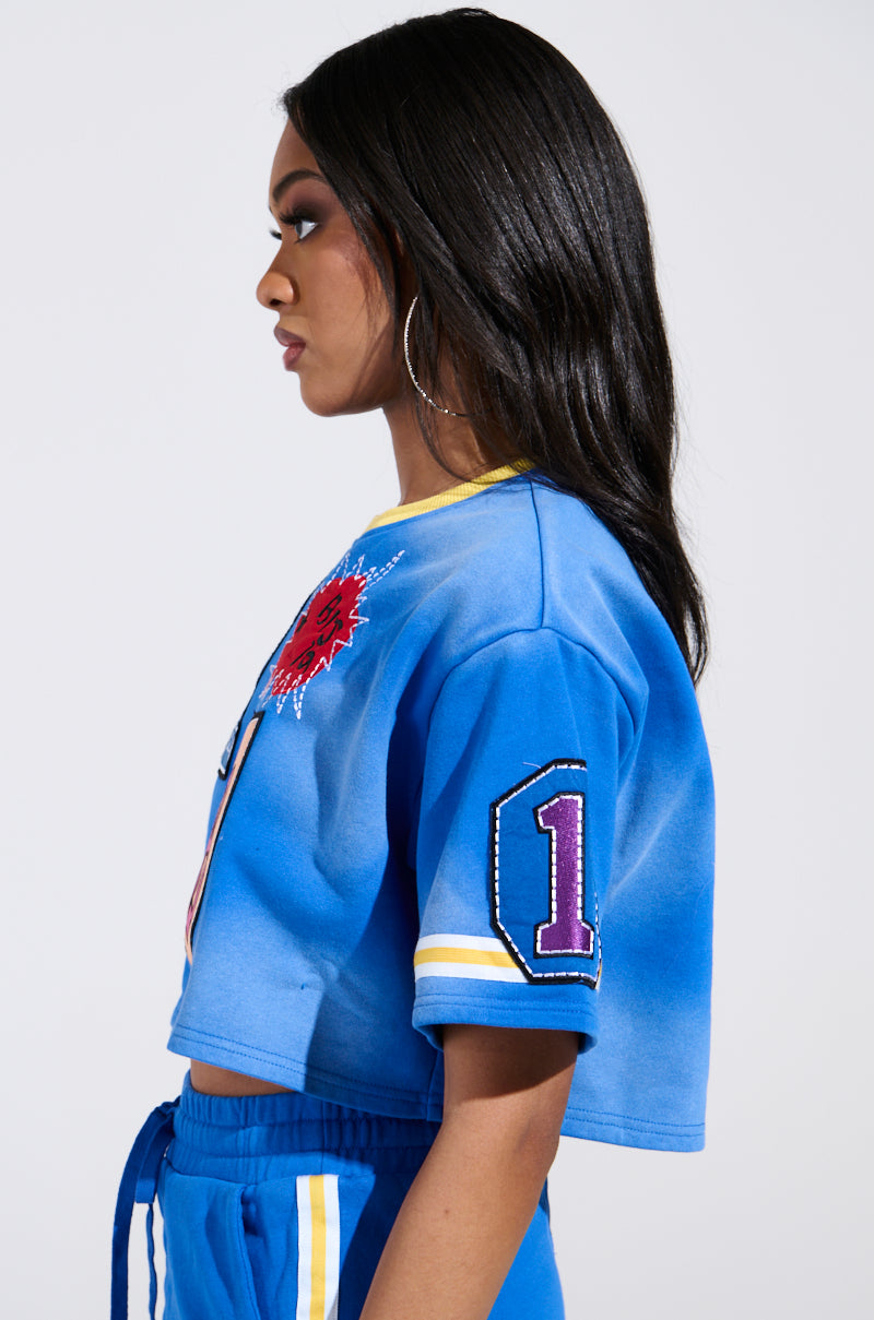 A Chicagoan with long dark hair is shown in profile, wearing a blue THREE ONE TWO CROP TEE by AKIRA Label and matching pants. The top features a red graphic and a decorated number "7" on the sleeve, as they look to the left.