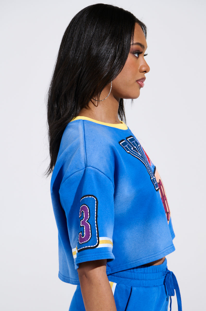 A Chicagoan with long black hair stands in profile against a plain background, wearing the AKIRA Label THREE ONE TWO CROP TEE. The blue crop tee has the number "3" on the sleeve and vibrant patterns on the front, complementing her large hoop earrings.