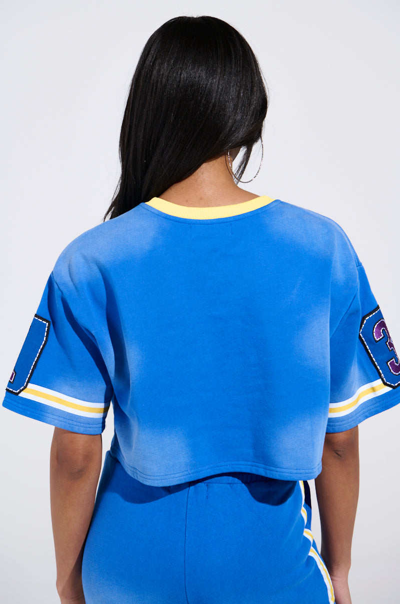 A Chicagoan with long black hair wears a blue cropped sports jersey, the THREE ONE TWO CROP TEE from the AKIRA Label, featuring yellow sleeve numbers. They pair it with matching blue pants, all set against a well-lit, light backdrop.