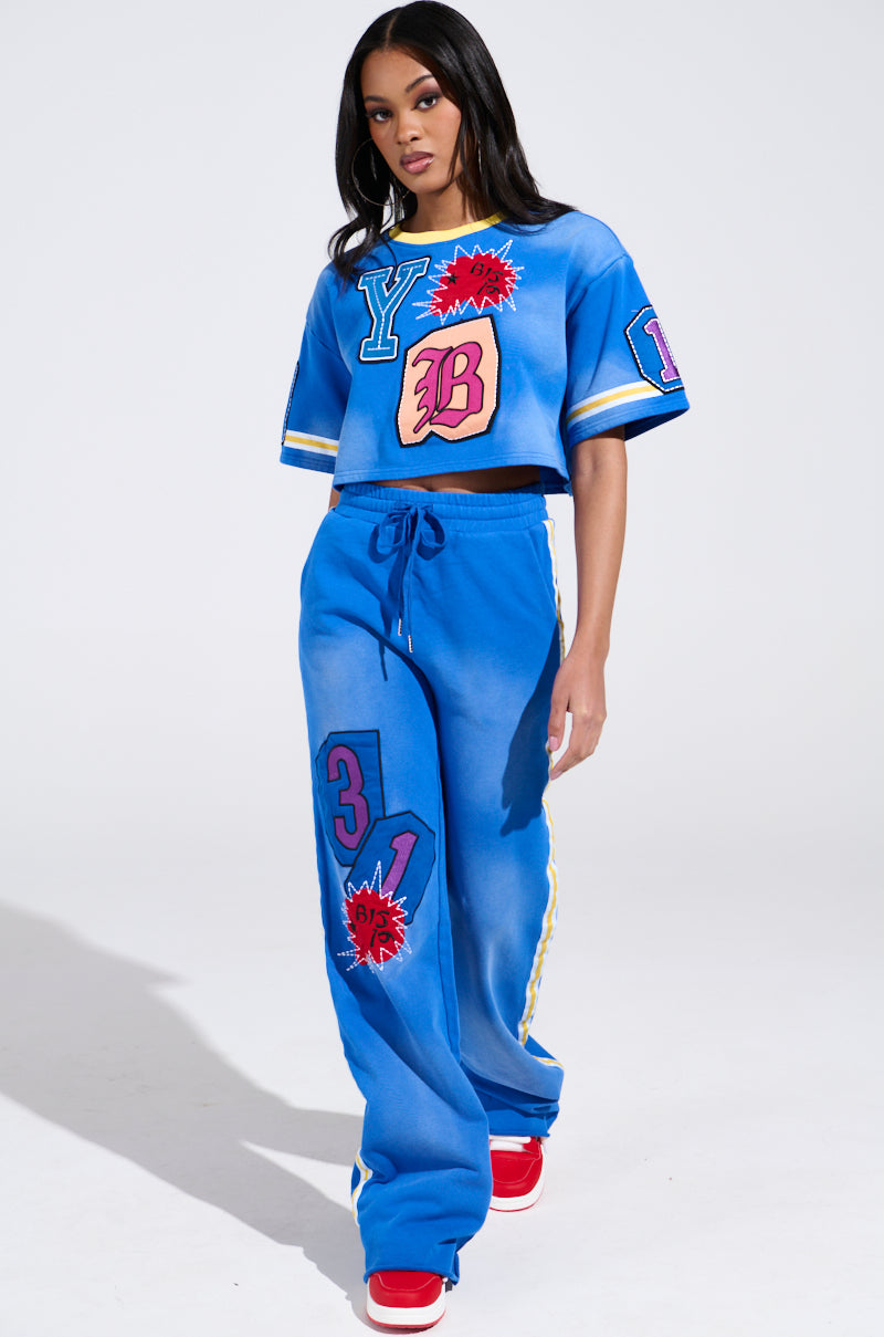 In Chicago, a woman confidently wears the AKIRA Label's THREE ONE TWO CROP TEE with matching vibrant blue pants, both decorated with colorful letter and number patches. She completes her look with red sneakers against a plain white background.