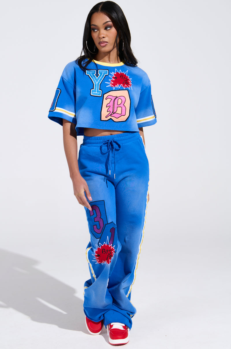 A Chicagoan woman wears an AKIRA Label THREE ONE TWO CROP TEE, showcasing graphic patches with yellow stripes and colorful comic-style designs. Her dynamic look is completed with matching pants and red sneakers.