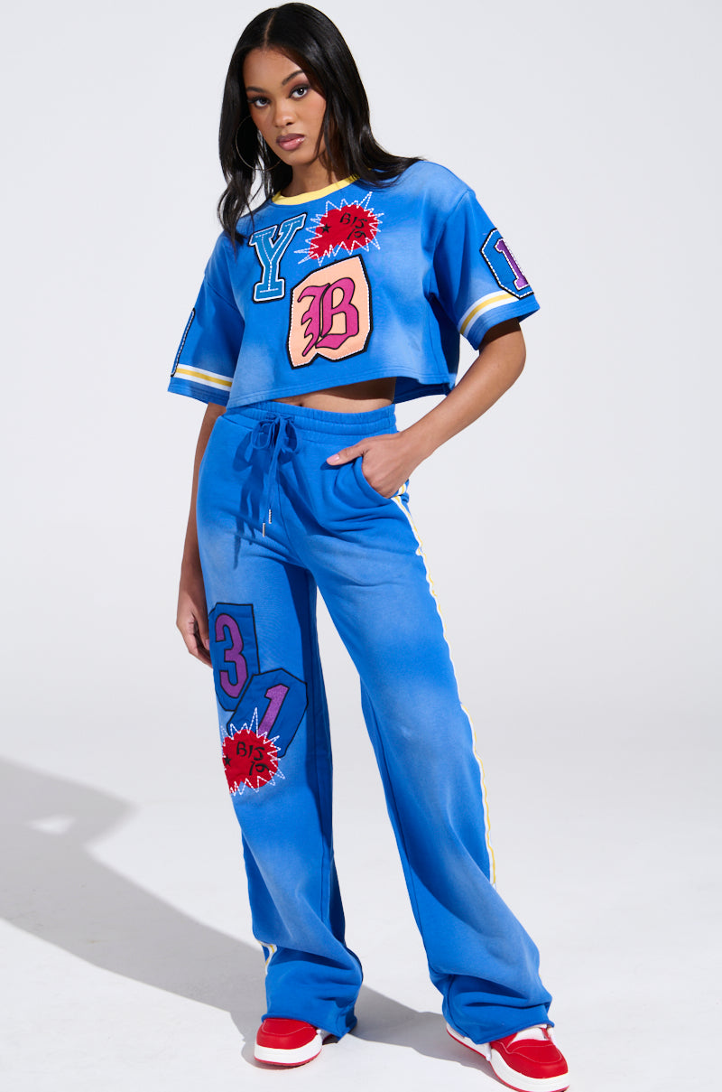 A Chicagoan strikes a pose in the THREE ONE TWO CROP TEE from the AKIRA Label, paired with vibrant blue track pants featuring bold graphic patches in red, yellow, and pink. Red shoes complete the look as their long, straight hair flows against a plain white background.
