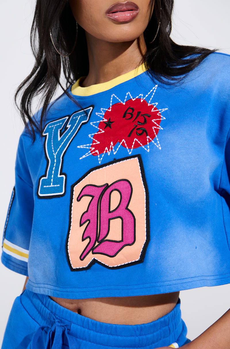 A person in a vibrant THREE ONE TWO CROP TEE featuring colorful "Y" and "B" patches and a red starburst, paired with blue wide-leg joggers, with long dark hair visible from the shoulders up.