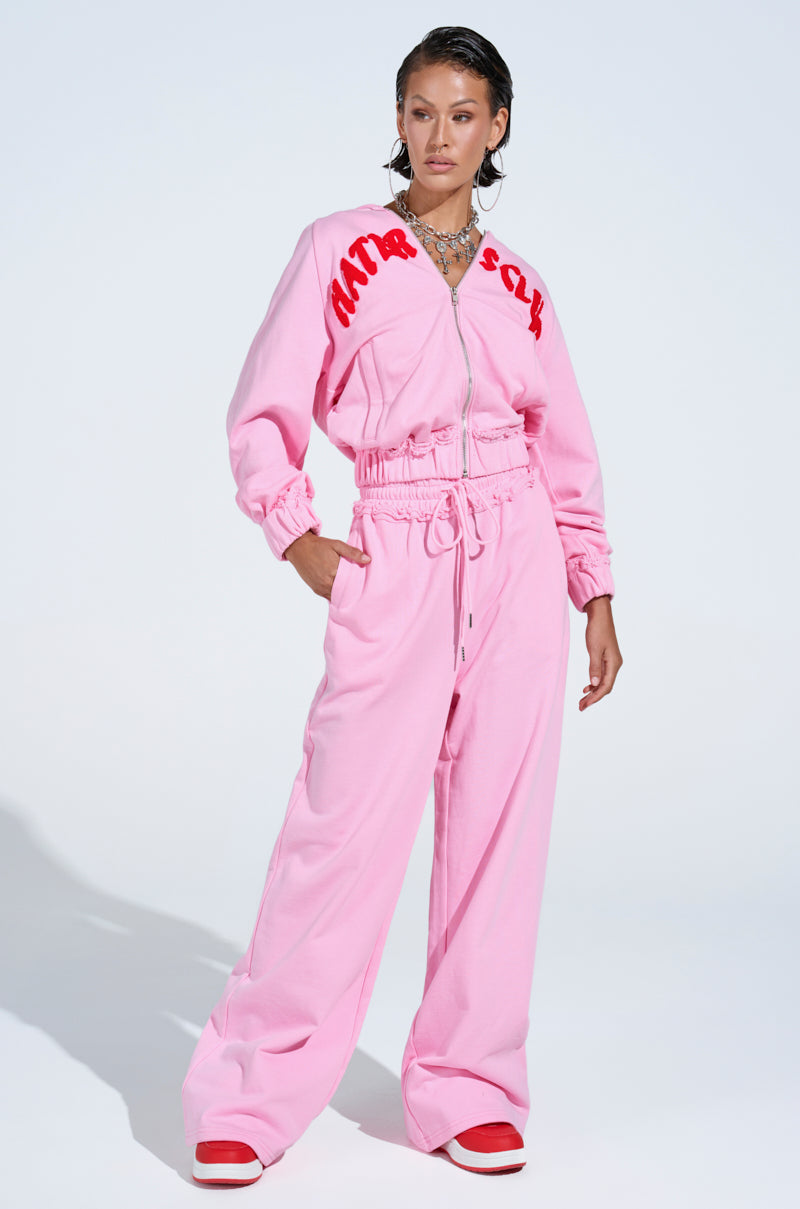 A woman stands confidently in AKIRA Label HATERS CLUB WIDE LEG JOGGER pants, styled with red and white sneakers, wet-look hair, and layered necklaces against a simple light background.