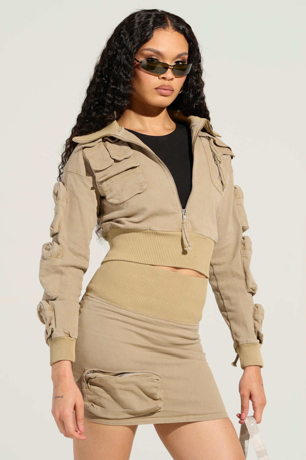 A woman with long curly hair wears sunglasses and sports the MY POCKETS DEEPER THAN YOURS BOMBER IN TAUPE, a beige jacket crafted from a stretchy cotton blend with pocket details. Her matching cargo-style skirt, featuring a pocket, enhances her ensemble. She carries a bag and stands against a plain light background.