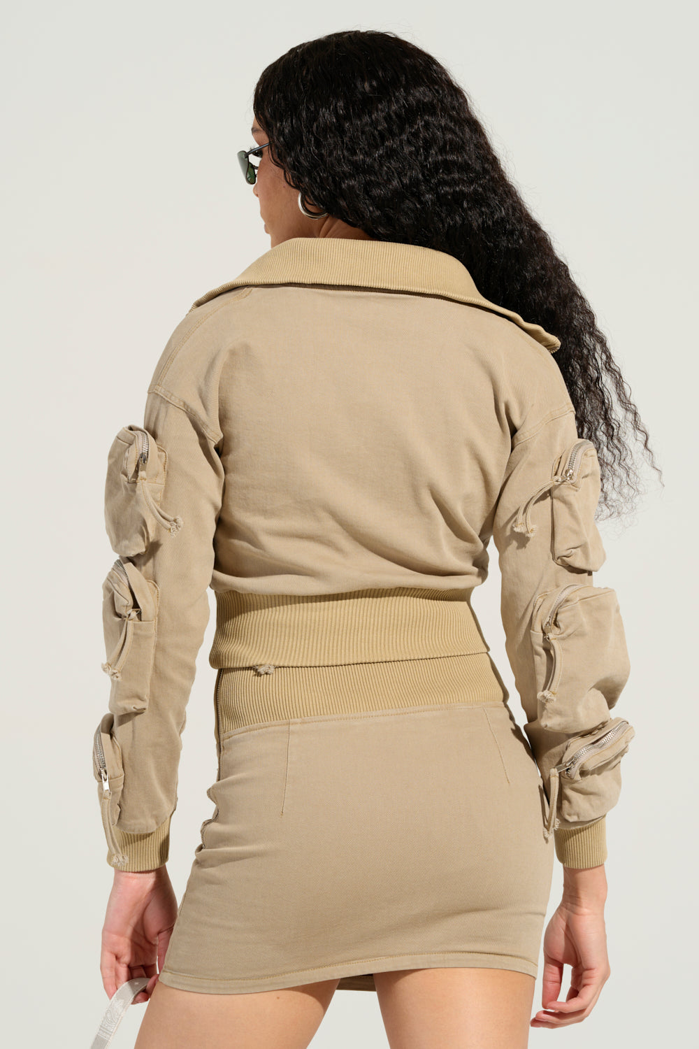 A person with long curly hair stands facing away, wearing a beige skirt and the "My Pockets Deeper Than Yours Bomber in Taupe," which features decorative zippered pouches on the sleeves. They are also wearing sunglasses against a plain, light background.