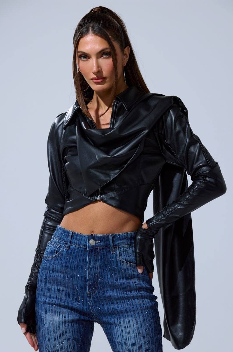 A woman with long brown hair wears the I'M NOT LIKE THE REST TOP—a shiny black faux leather cropped jacket with draped details—paired with fingerless gloves and high-waisted blue jeans, against a plain gray background.