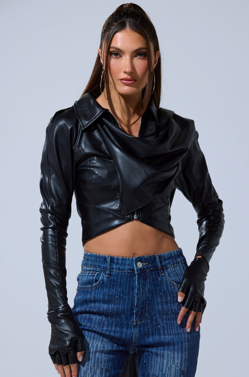 A woman wears the I'M NOT LIKE THE REST TOP—a black faux leather cropped jacket with strappy details—paired with fingerless gloves and blue high-waisted jeans, standing against a plain light background.