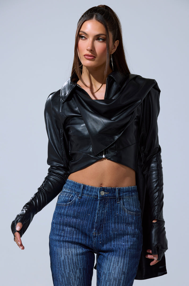 A woman with long brown hair wears the I'M NOT LIKE THE REST TOP, a black faux leather cropped jacket with strappy details and fingerless gloves, paired with high-waisted blue jeans. She poses against a plain light gray background.