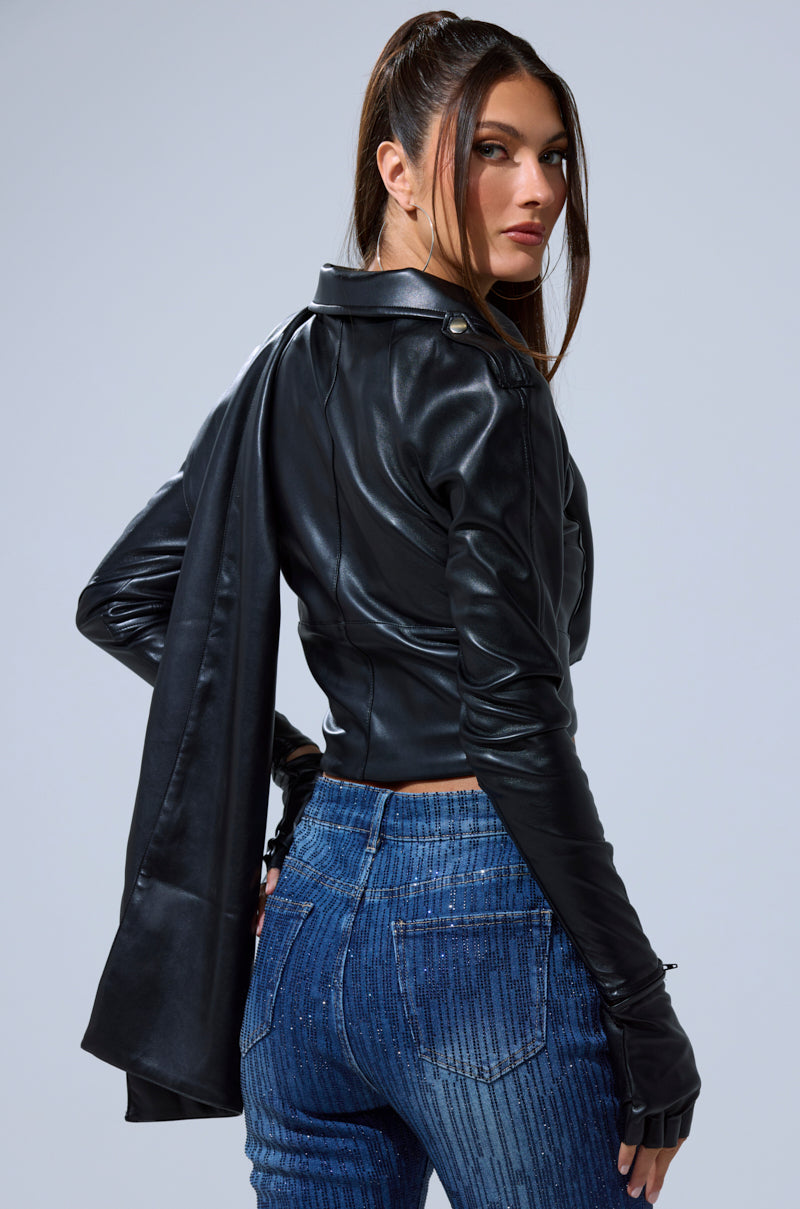 A woman with long brown hair in a ponytail wears the I'M NOT LIKE THE REST TOP, blue high-waisted jeans with vertical lines, and black gloves. She looks over her shoulder against a plain light gray background.