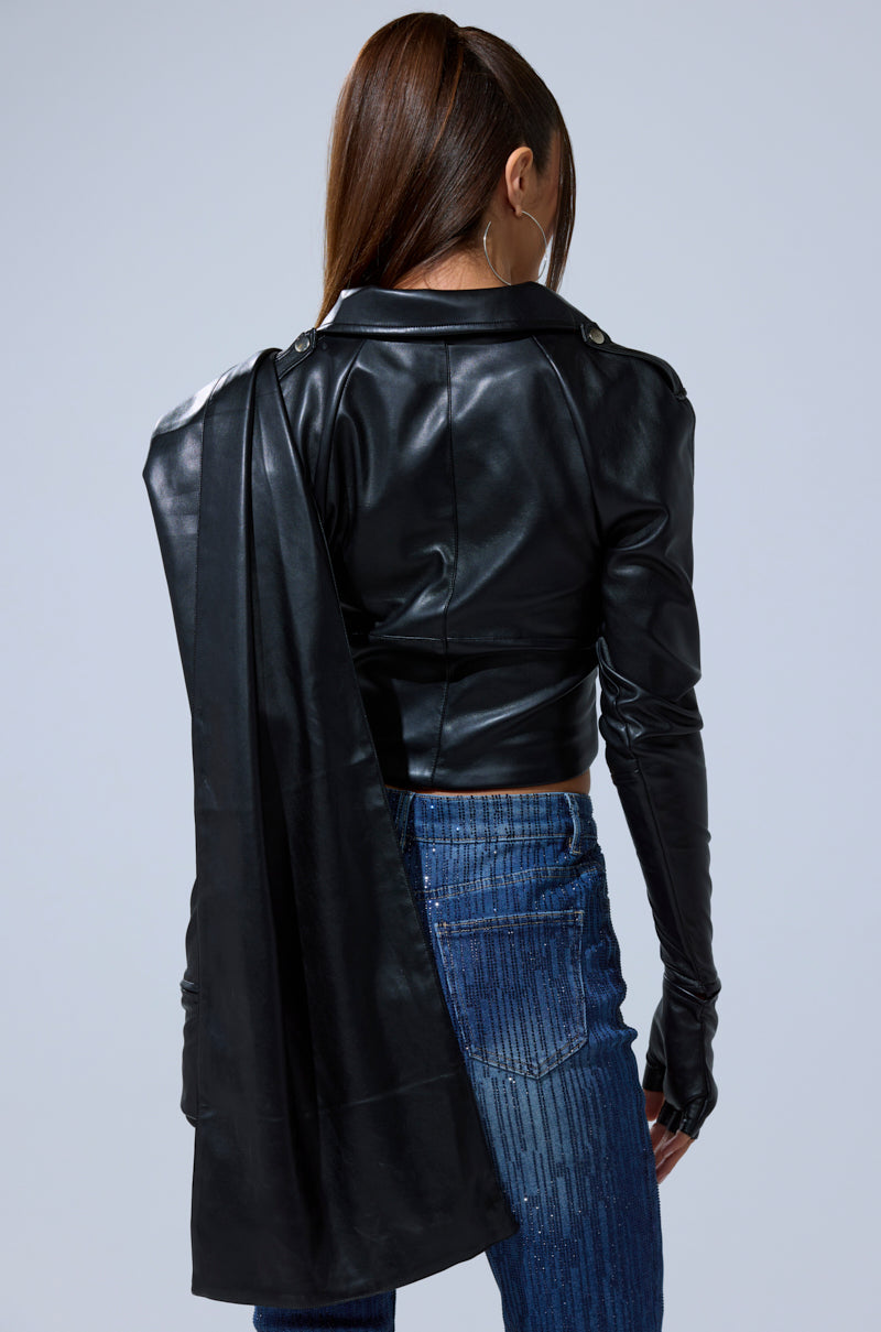 A woman with long brown hair in a ponytail wears the I'M NOT LIKE THE REST TOP, featuring strappy black faux leather detail, paired with blue jeans. She stands with her back to the camera, a black scarf over her left shoulder.
