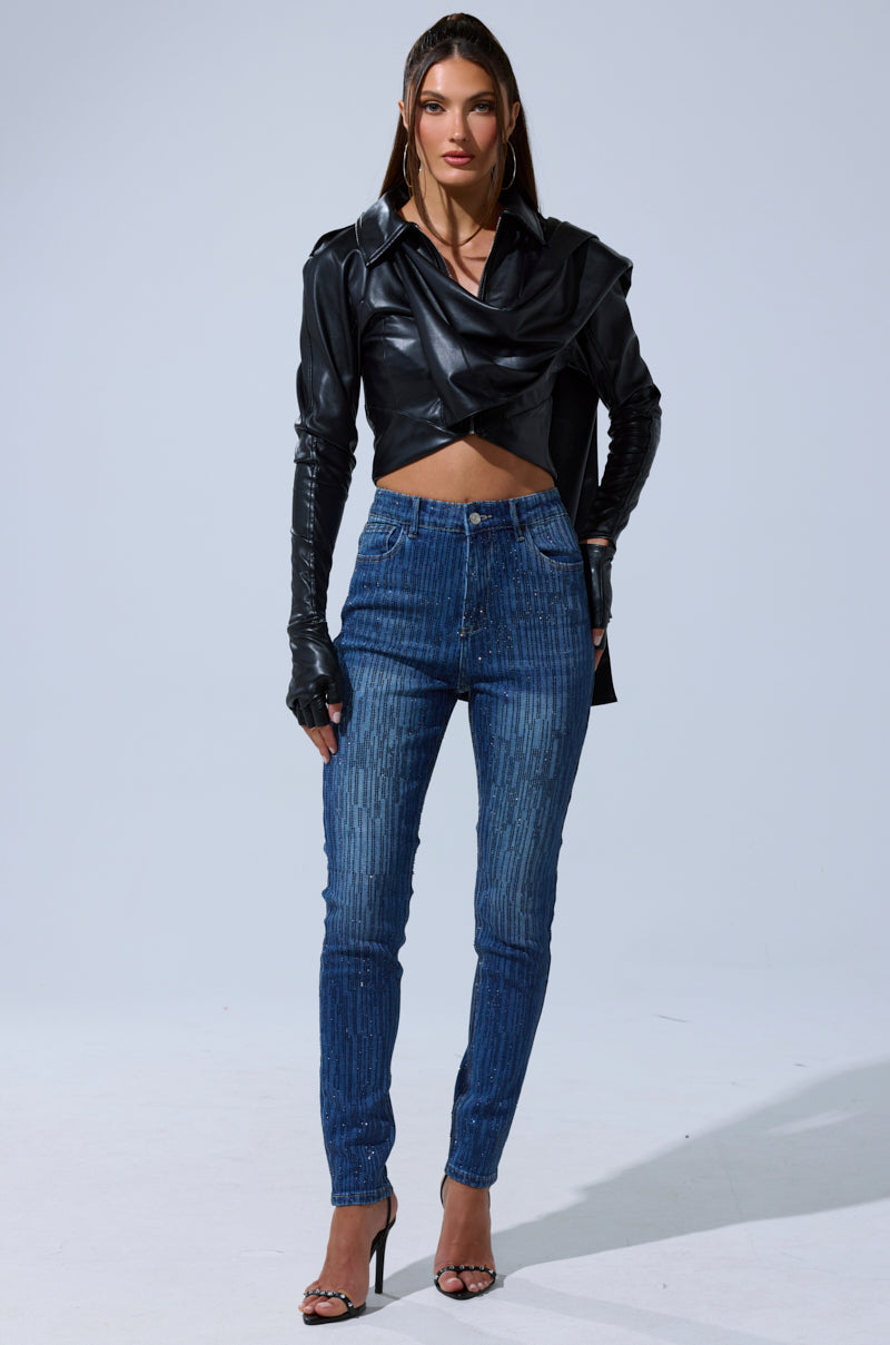 A woman faces forward, wearing the I'M NOT LIKE THE REST TOP— a black draped faux leather piece—paired with blue patterned jeans, black high-heeled sandals, and gloves. Her straight hair is tucked behind her ears as she poses against a light background.