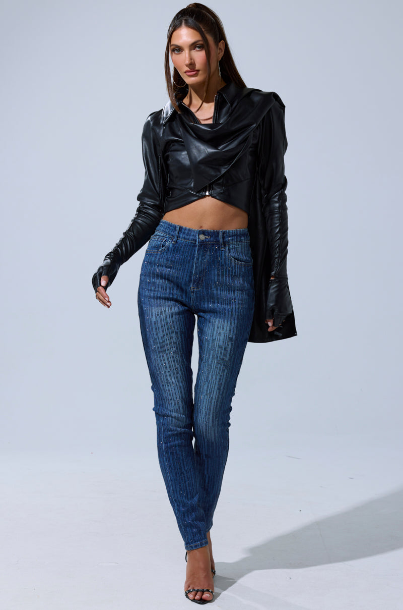 A woman stands confidently in the I'M NOT LIKE THE REST TOP, a black asymmetrical faux leather piece with a draped silhouette, paired with blue high-waisted skinny jeans and black high-heeled sandals against a light gray background.