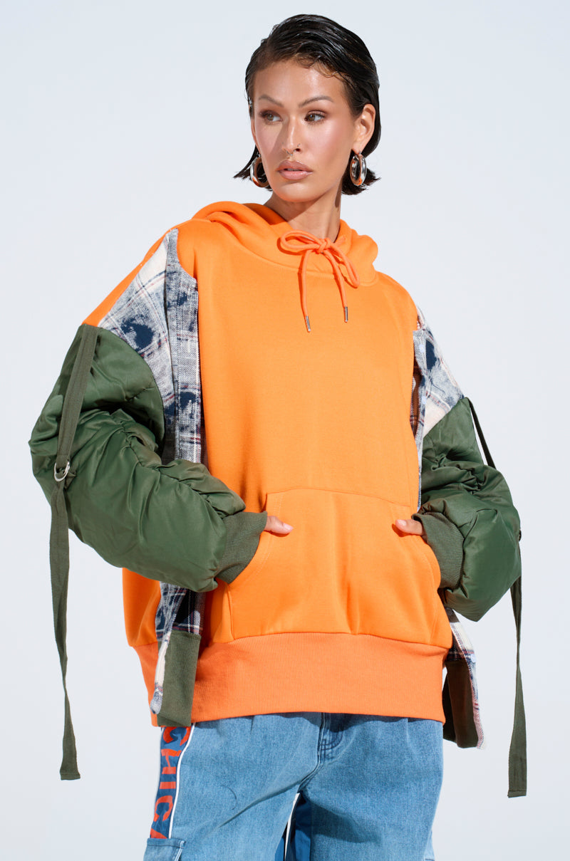 Wearing the MY EX IS MY BIGGEST FAN REBUILT SWEATSHIRT—an oversized orange hoodie with green puffer sleeves and patterned details—a woman pairs it with blue jeans featuring orange lettering, standing hands-in-pocket against a plain background. 1