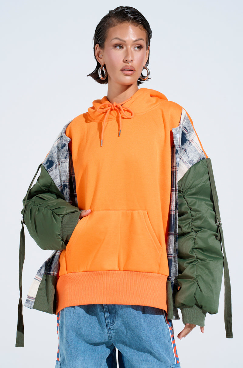 A woman wears the MY EX IS MY BIGGEST FAN REBUILT SWEATSHIRT, an oversized bright orange hoodie under a patchwork jacket with green sleeves and plaid details, styled with blue jeans, short wet-look hair, large hoop earrings, against a plain background. 2