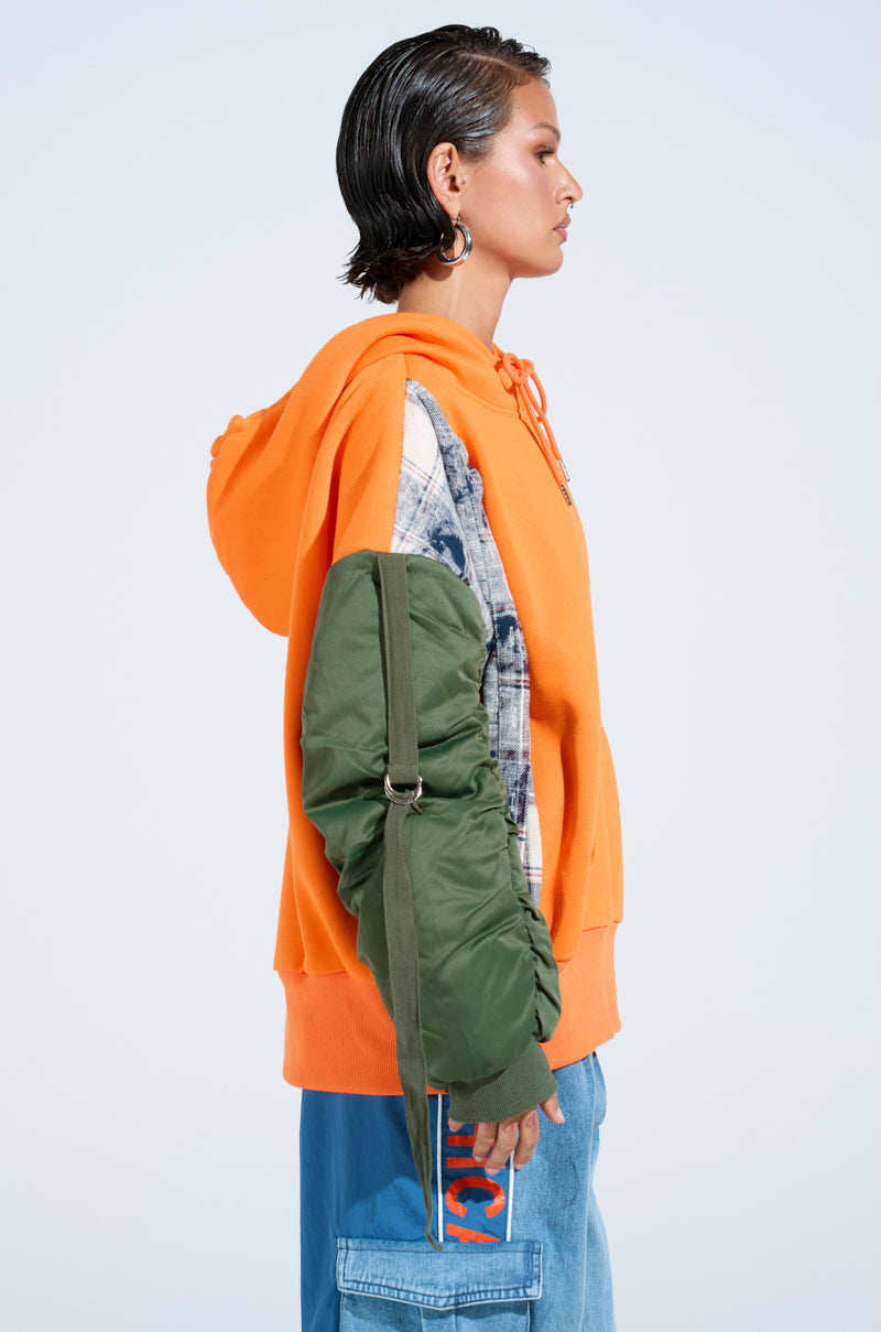 A person with short, slicked-back hair wears the oversized MY EX IS MY BIGGEST FAN REBUILT SWEATSHIRT in orange with patchwork sleeves, blue jeans, hoop earrings, and a green strap bag, standing in profile against a plain background. 3