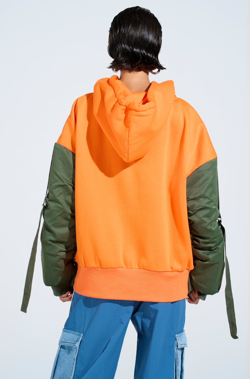 A person stands with their back to the camera, wearing an oversized MY EX IS MY BIGGEST FAN REBUILT SWEATSHIRT—an orange hoodie with green sleeves and long straps—paired with blue cargo pants against a plain light background. 4