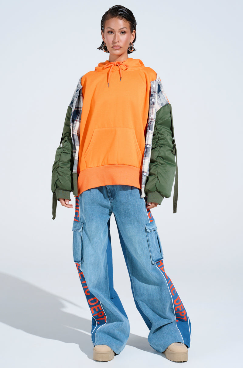 Wearing the MY EX IS MY BIGGEST FAN REBUILT SWEATSHIRT in orange, a mixed-material green and plaid jacket, wide-leg blue cargo jeans with red side text, and beige shoes, a person faces forward against a light backdrop in an oversized look. 5