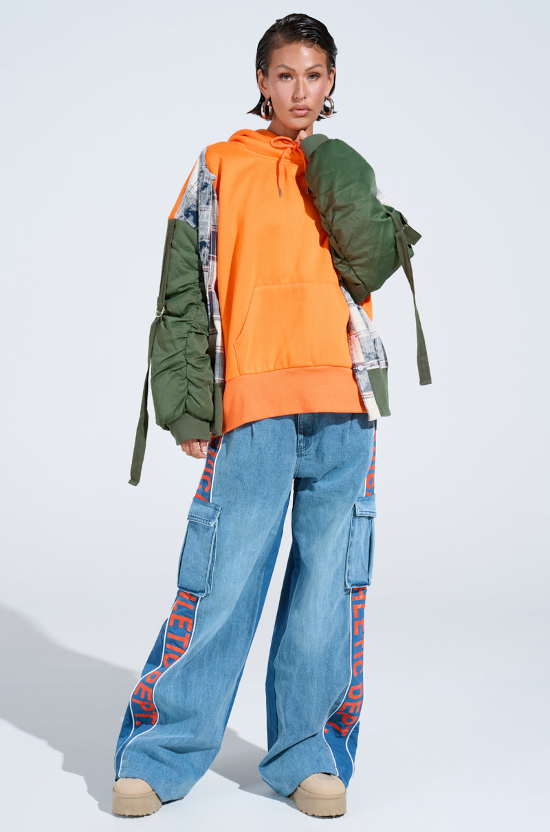 A person poses in the MY EX IS MY BIGGEST FAN REBUILT SWEATSHIRT with green quilted sleeves, oversized blue cargo jeans featuring red text, beige platform shoes, and hoop earrings against a plain background. 6