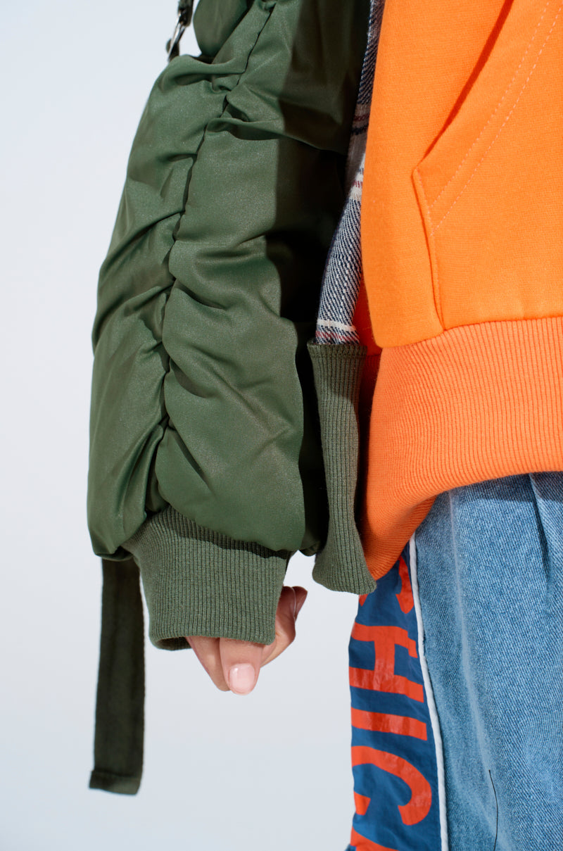 A close-up shows a person wearing the MY EX IS MY BIGGEST FAN REBUILT SWEATSHIRT in orange, paired with a green ruched sleeve jacket and blue jeans featuring red text. Only their hand and part of the outfit are visible. 7
