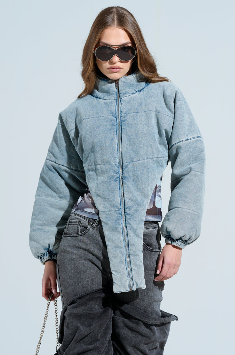 A person with long brown hair poses coolly against a light blue background, wearing oversized sunglasses and the CHARLI CROPPED DENIM PUFFER. They're holding a chain-strap handbag, exuding confidence.