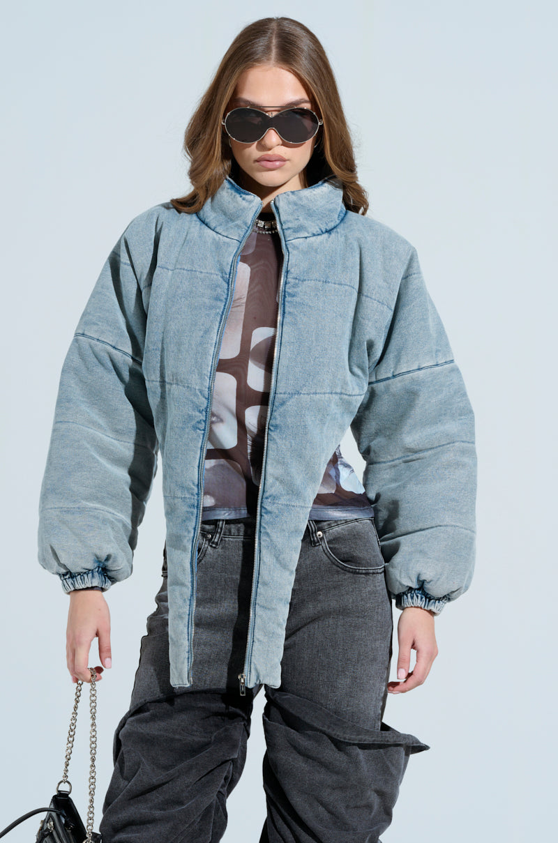 A woman with long brown hair and large black sunglasses confidently stands against a light background, wearing a CHARLI CROPPED DENIM PUFFER and dark grey pants, holding a black purse in her right hand—a chic blend of style and practicality for cold-weather elegance.