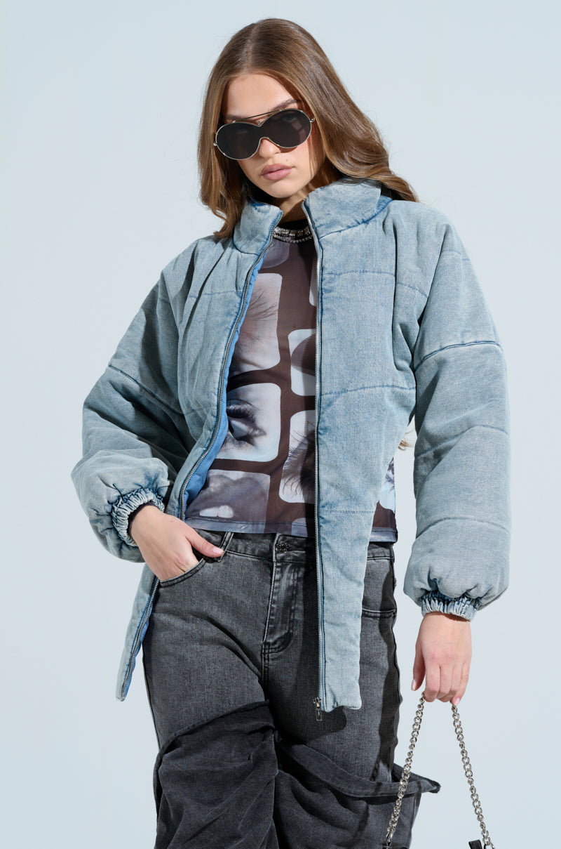 A person sporting a CHARLI CROPPED DENIM PUFFER, graphic tee, and dark jeans carries a chain handbag. With long hair and sunglasses, they pose casually against a light blue backdrop.
