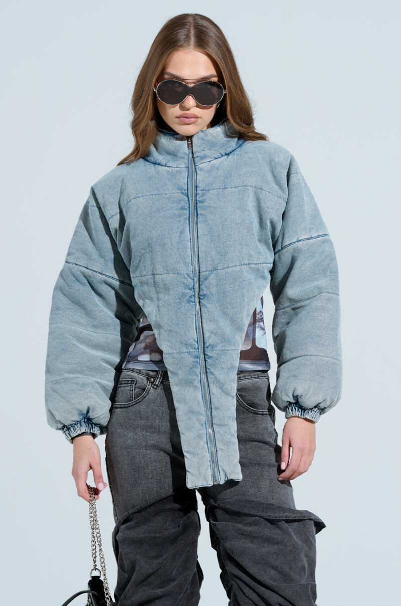 Wearing oversized sunglasses and the CHARLI CROPPED DENIM PUFFER, someone stands confidently against a plain background with a chain-strap bag and dark gray pants, exuding fashion-forward style.