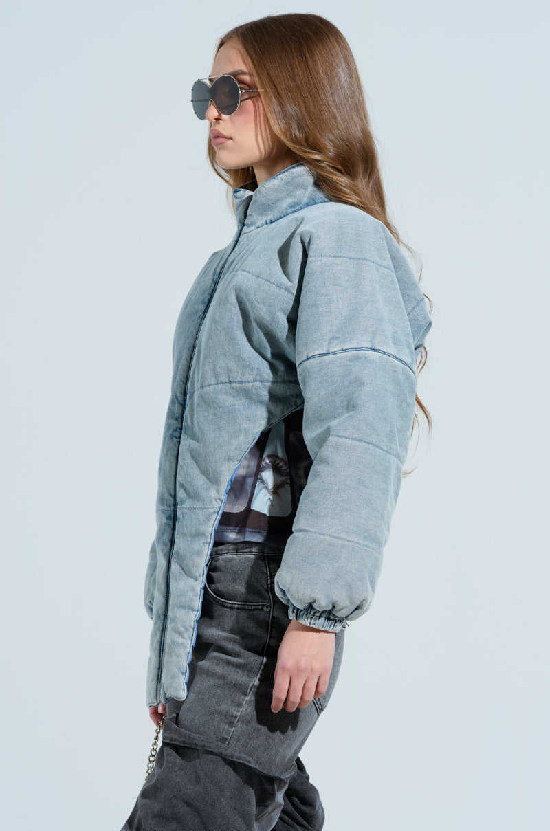 A person with long brown hair and sunglasses stands against a light background, wearing the CHARLI CROPPED DENIM PUFFER and dark pants, effortlessly showcasing perfect cold-weather style.