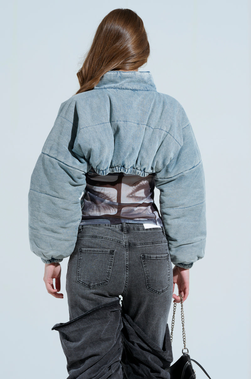 A person with long hair faces away in a chic CHARLI CROPPED DENIM PUFFER and high-waisted dark gray jeans, holding a small chain-strap purse against a plain light background.