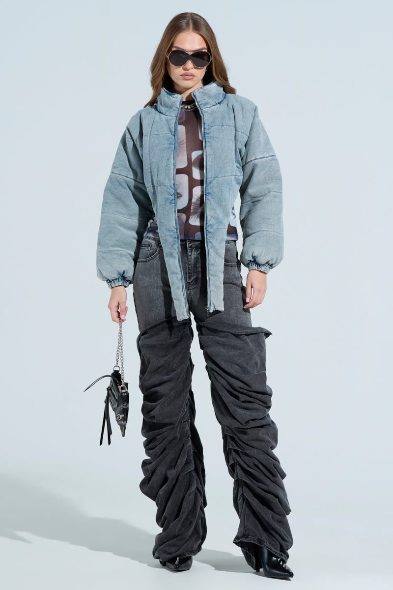 In oversized dark gray ruched pants, a gray padded jacket, and a graphic T-shirt with a small handbag, they wear sunglasses over long brown hair. The outfit features the CHARLI CROPPED DENIM PUFFER against a plain light background.