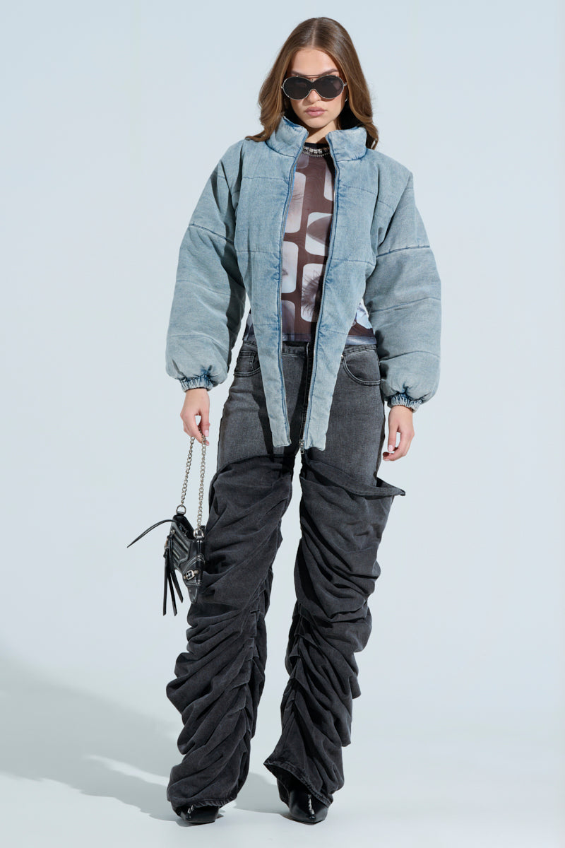 Sporting oversized ruched black pants and a printed shirt, an individual in a CHARLI CROPPED DENIM PUFFER poses against a plain backdrop. They have long hair and large sunglasses, holding a small black purse with a chain strap.